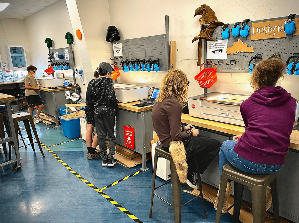 Several students use Glowforge laser engravers in a makerspace equipped with safety gear and tools.