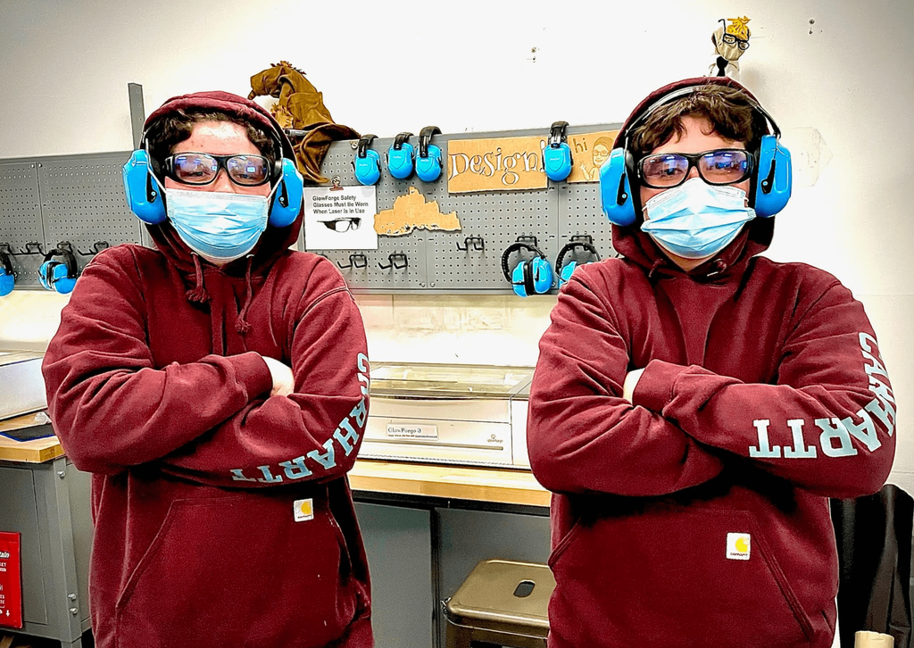 Two students wearing matching maroon hoodies, blue earmuffs, and masks pose confidently in the makerspace.