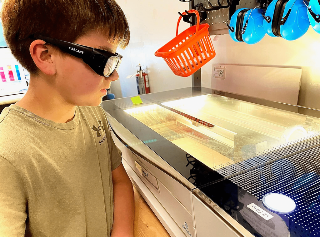 A student in a tan shirt and safety glasses monitors a laser engraving project inside the Glowforge machine.