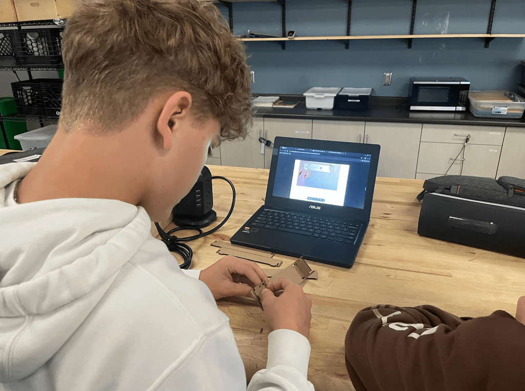 A student works at a wooden table assembling a small cardboard project while following instructions on a laptop.