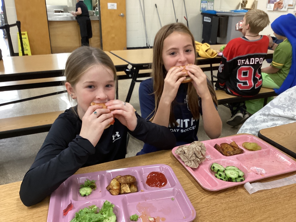 Two students sit at a cafeteria table enjoying their sandwiches and smiling, with pink trays of lunch food including vegetables and tater tots in front of them.