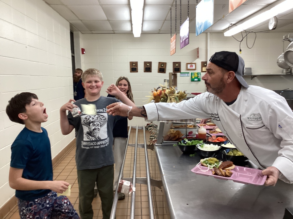 Chef Tony playfully tosses a slice of cheese toward a laughing student while others in line smile and cheer, creating a lively, fun cafeteria moment.