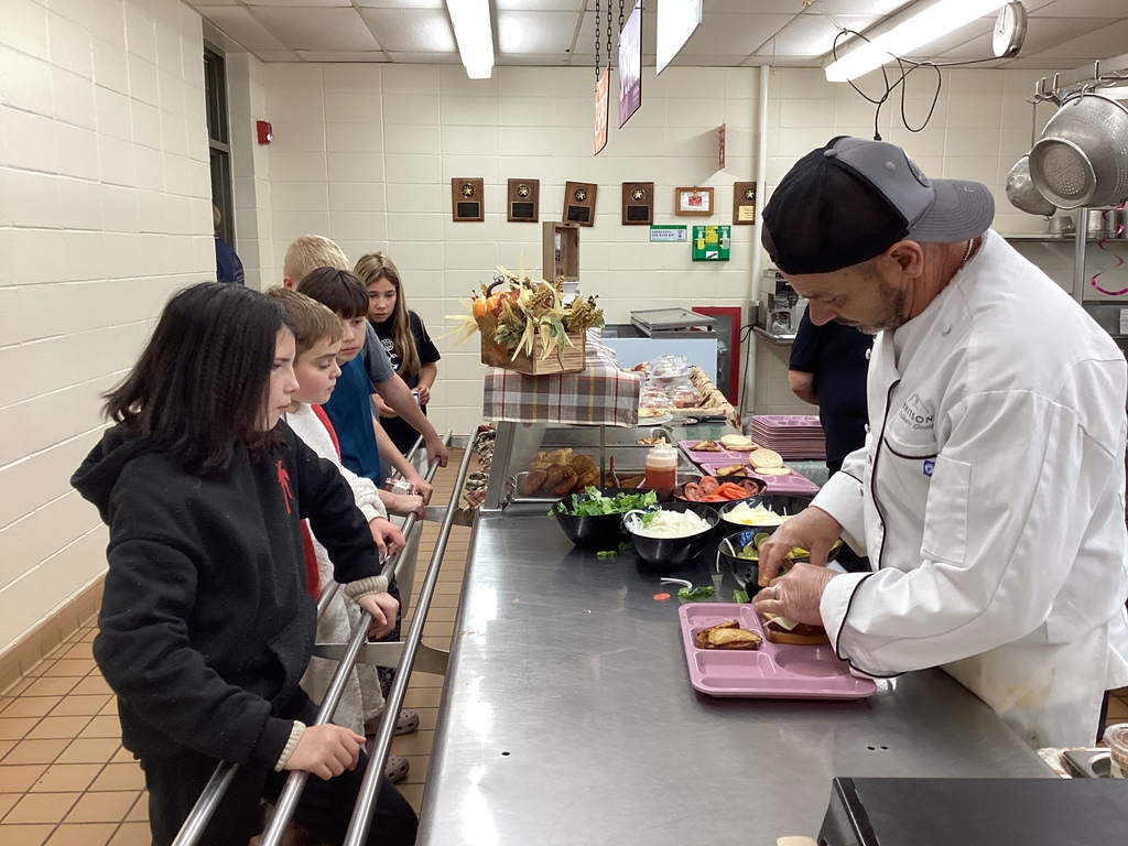 Chef Tony prepares sandwiches behind the counter while a group of students eagerly waits, watching him assemble their lunches.