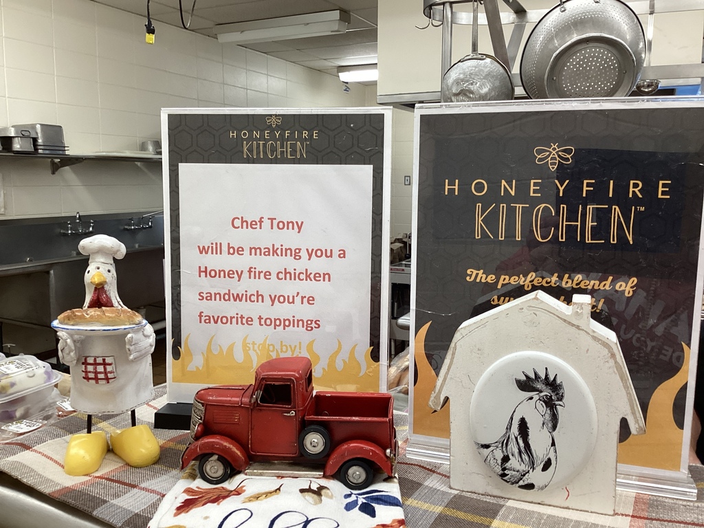 A Honeyfire Kitchen display featuring a sign that reads “Chef Tony will be making you a Honey fire chicken sandwich with your favorite toppings,” surrounded by fall-themed decorations, a small red toy truck, and chicken figurines.
