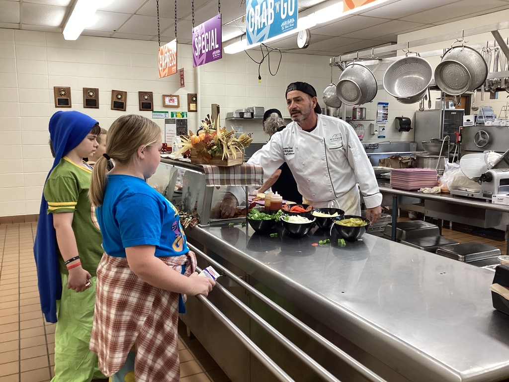 Chef Tony, wearing a white chef’s coat and black cap, talks with two elementary students dressed in colorful outfits at the lunch counter, with bowls of sandwich toppings in front of him.