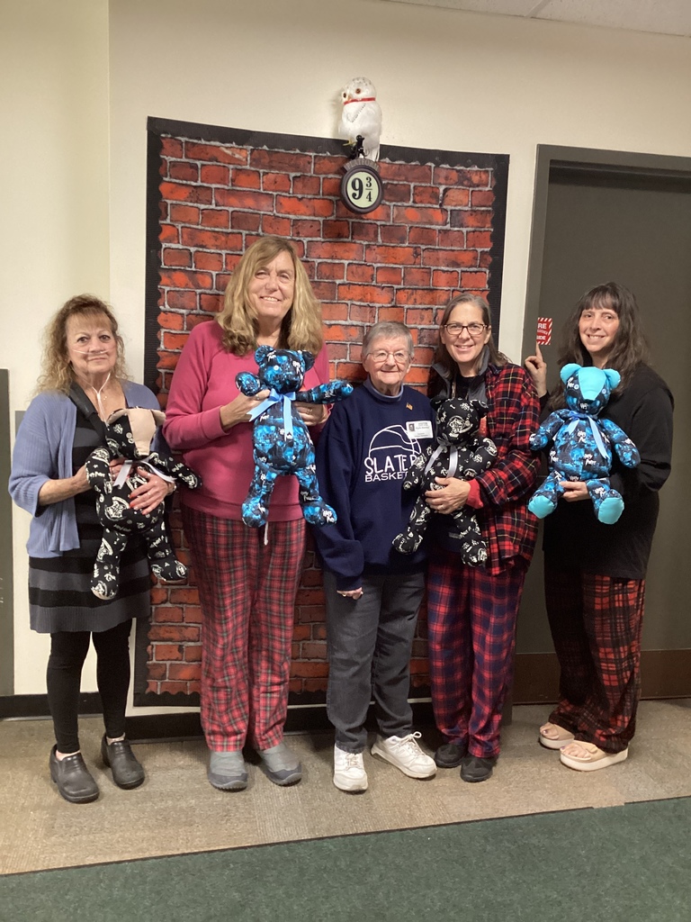 Five women dressed in plaid pajamas stand smiling in front of a brick-patterned backdrop with a “Platform 9¾” sign and a white owl decoration, each holding a handmade teddy bear.