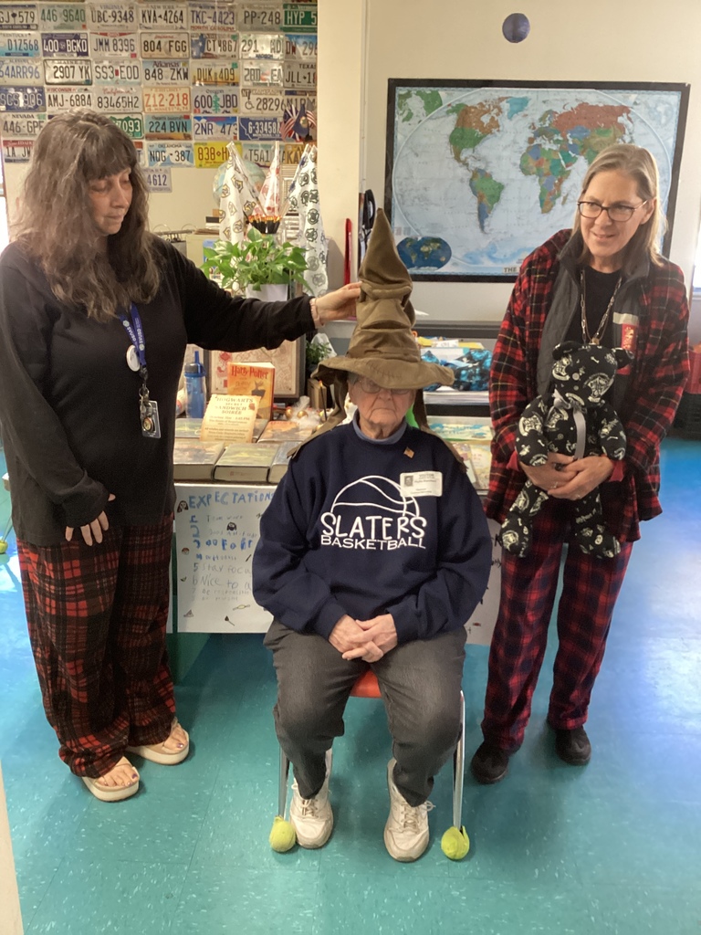 Two women stand beside a seated older woman wearing a “Slaters Basketball” sweatshirt and the Sorting Hat from Harry Potter, participating in a themed classroom activity.