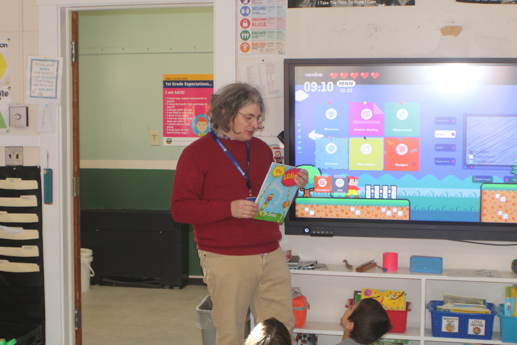 A teacher wearing a red sweater reads The Lorax by Dr. Seuss to a group of children seated on the floor, with a colorful classroom screen behind them.