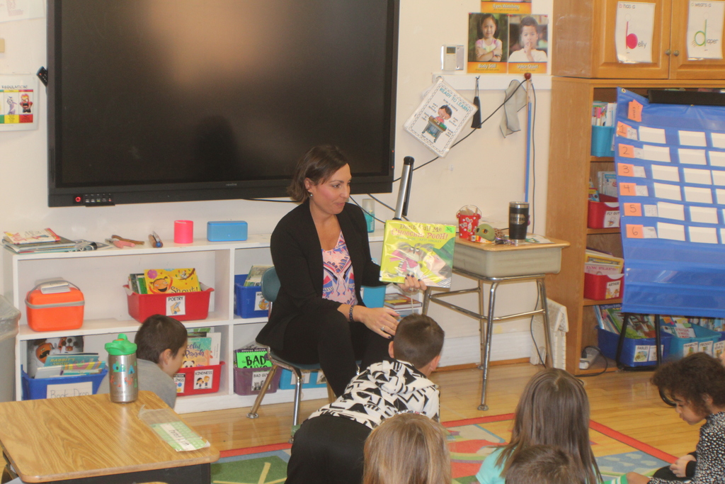 A woman sits in front of a classroom of young students, holding up a picture book while the children sit on a colorful rug listening attentively.
