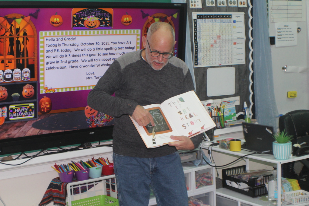 The same man reads a book to students with a Halloween-themed digital display in the background, featuring pumpkins and a classroom message.