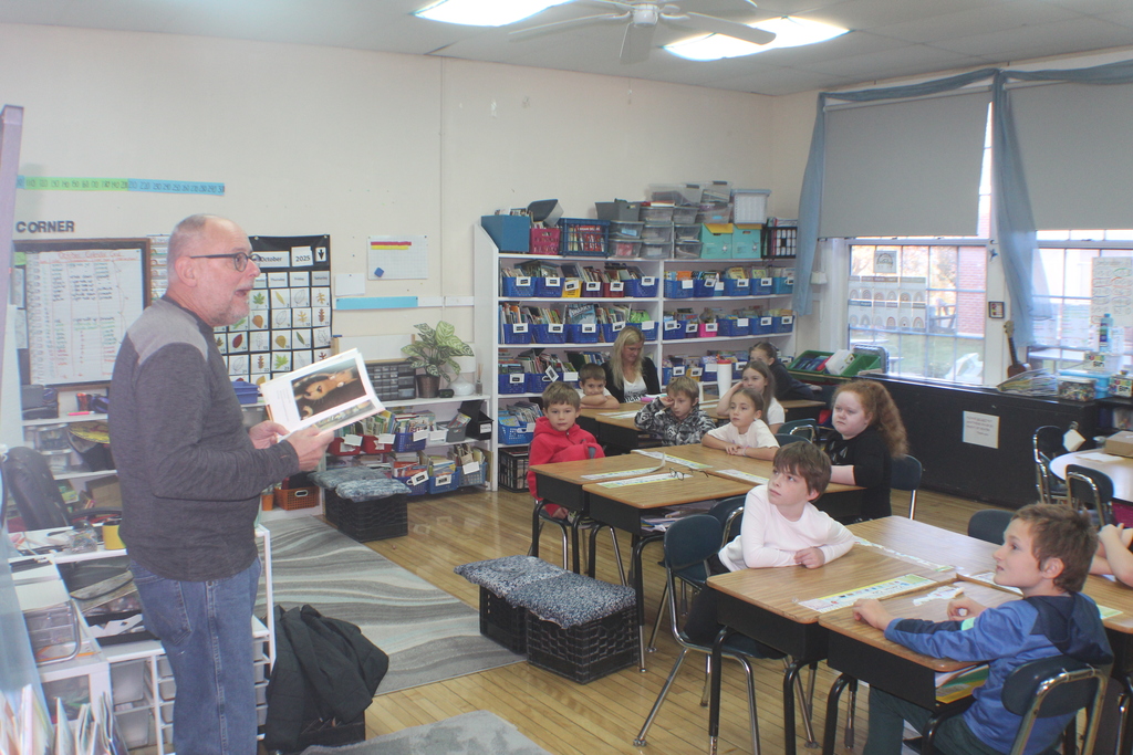 A man stands at the front of a classroom reading aloud to a group of elementary students seated at their desks, surrounded by shelves of books and classroom materials.