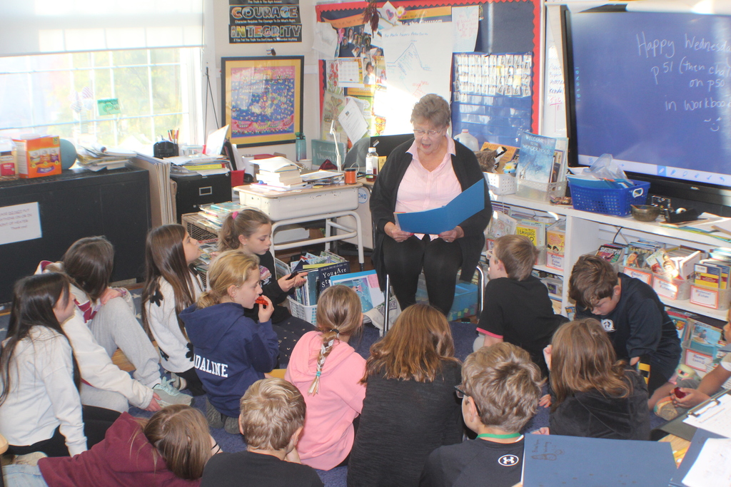 An older woman reads aloud to a group of attentive students sitting on the floor in a cozy, sunlit classroom filled with books and learning posters.