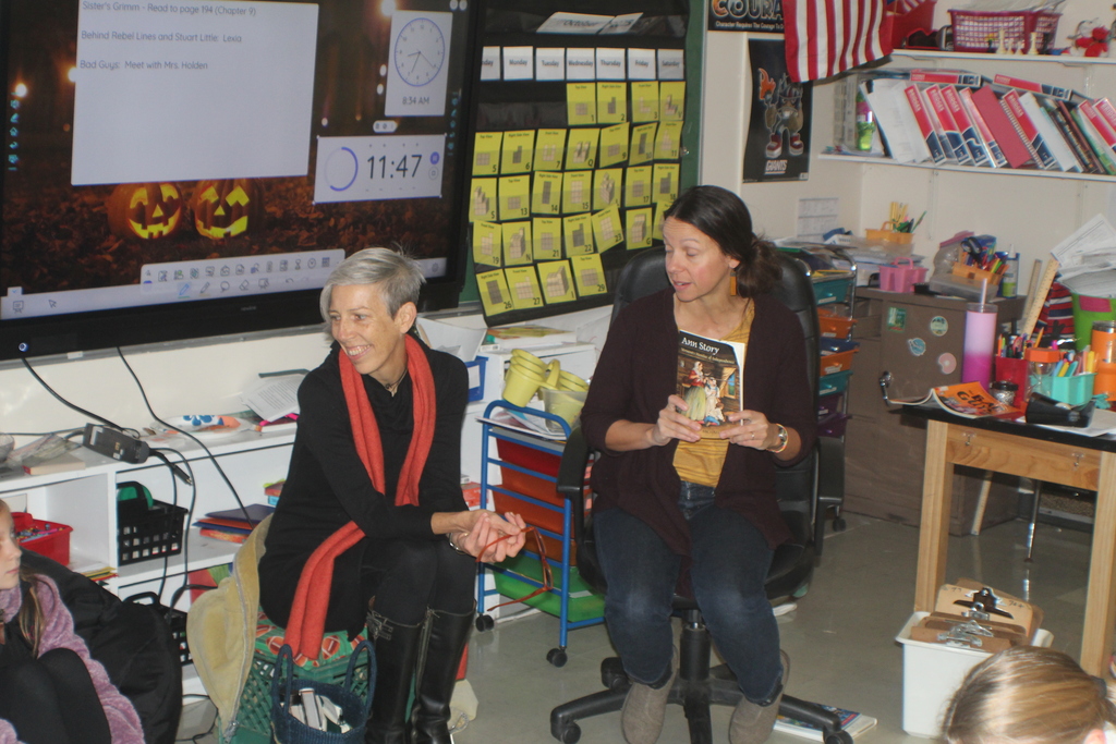 Two women sit at the front of a classroom; one holds a book titled Ann Story while the other engages with students during a lesson.