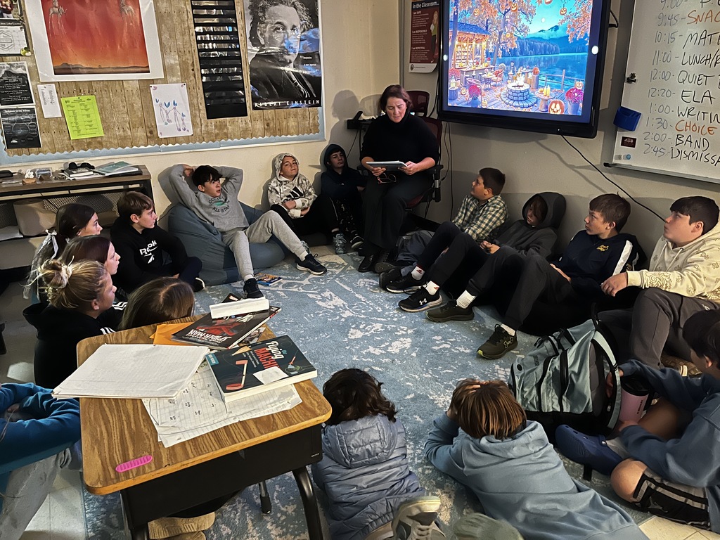 An adult reads aloud to a group of middle school students seated in a circle on the classroom floor, surrounded by books and notebooks, with a cozy fall-themed scene displayed on the screen behind them.