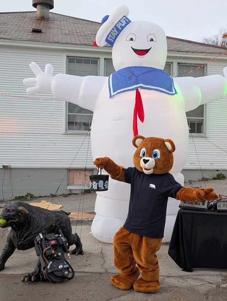 A person in a brown bear mascot costume holds a Halloween candy bucket and poses in front of a large inflatable Stay Puft Marshmallow Man decoration outside a building.