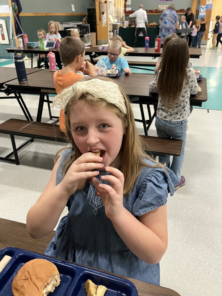 A student wearing a blue dress and headband eats lunch in the cafeteria, with other students seated in the background.