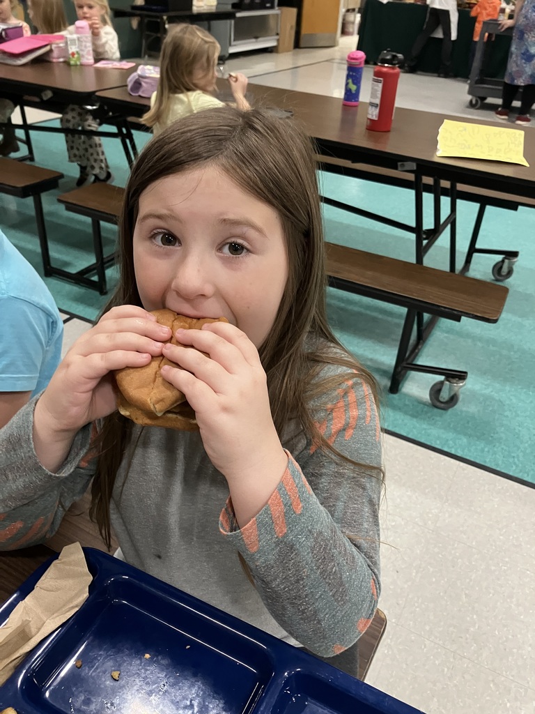 A student with long brown hair takes a bite of a sandwich at the cafeteria table.