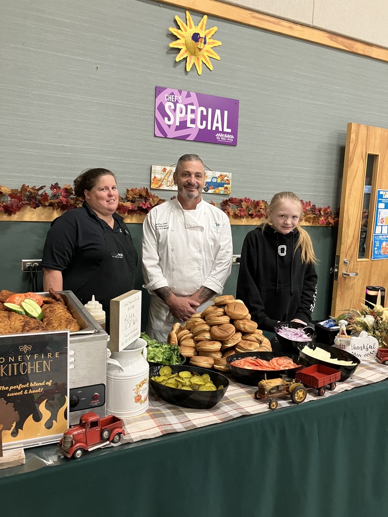 Two cafeteria staff members and a student stand behind a serving table filled with buns, vegetables, and chicken under a “Chef’s Special” sign.