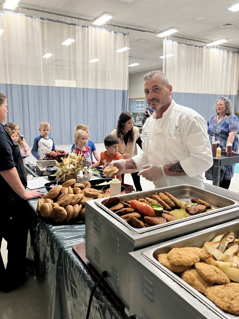 A chef prepares food behind a buffet line while students and staff wait to be served in the cafeteria.