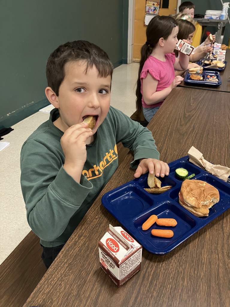 A student in a green sweatshirt eats a potato wedge at lunch with carrots, a sandwich, and chocolate milk on a blue tray.
