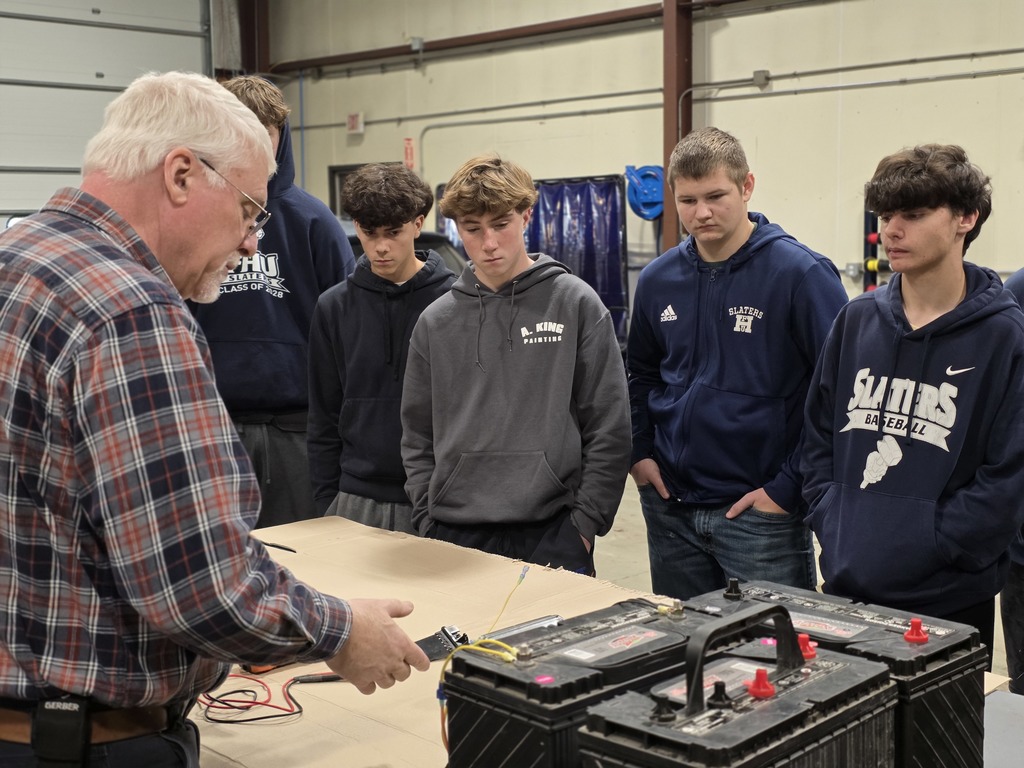A teacher demonstrates electrical testing with large truck batteries as several students watch attentively.