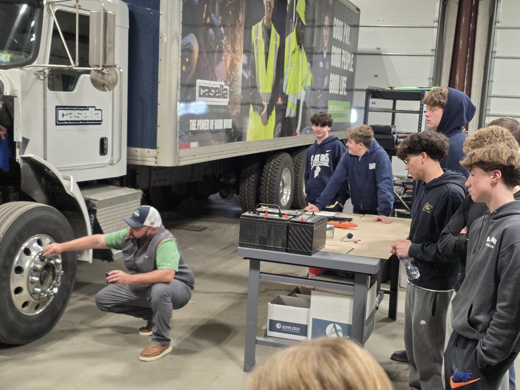 A Casella employee kneels beside a truck, explaining tire maintenance to students standing nearby.