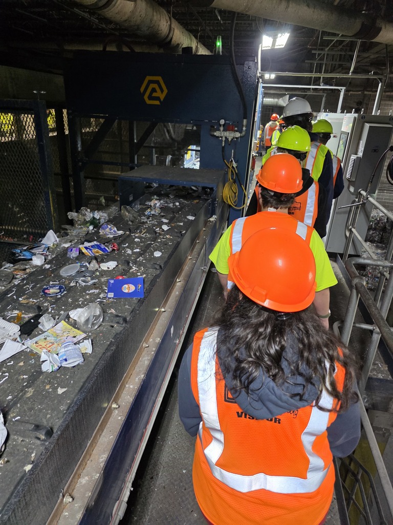 A line of students wearing orange and yellow safety vests walks beside a conveyor belt filled with recyclable items inside the plant.