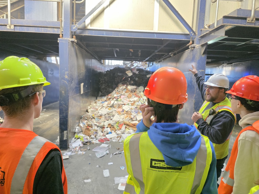 Students in safety gear listen as a tour guide gestures toward a large pile of recyclable materials inside a recycling facility.