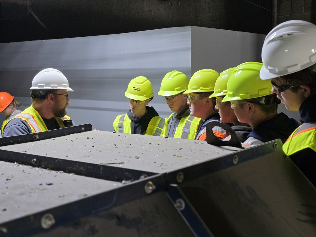 A staff member in a white hard hat explains machinery operations to a group of students in green helmets and safety vests.