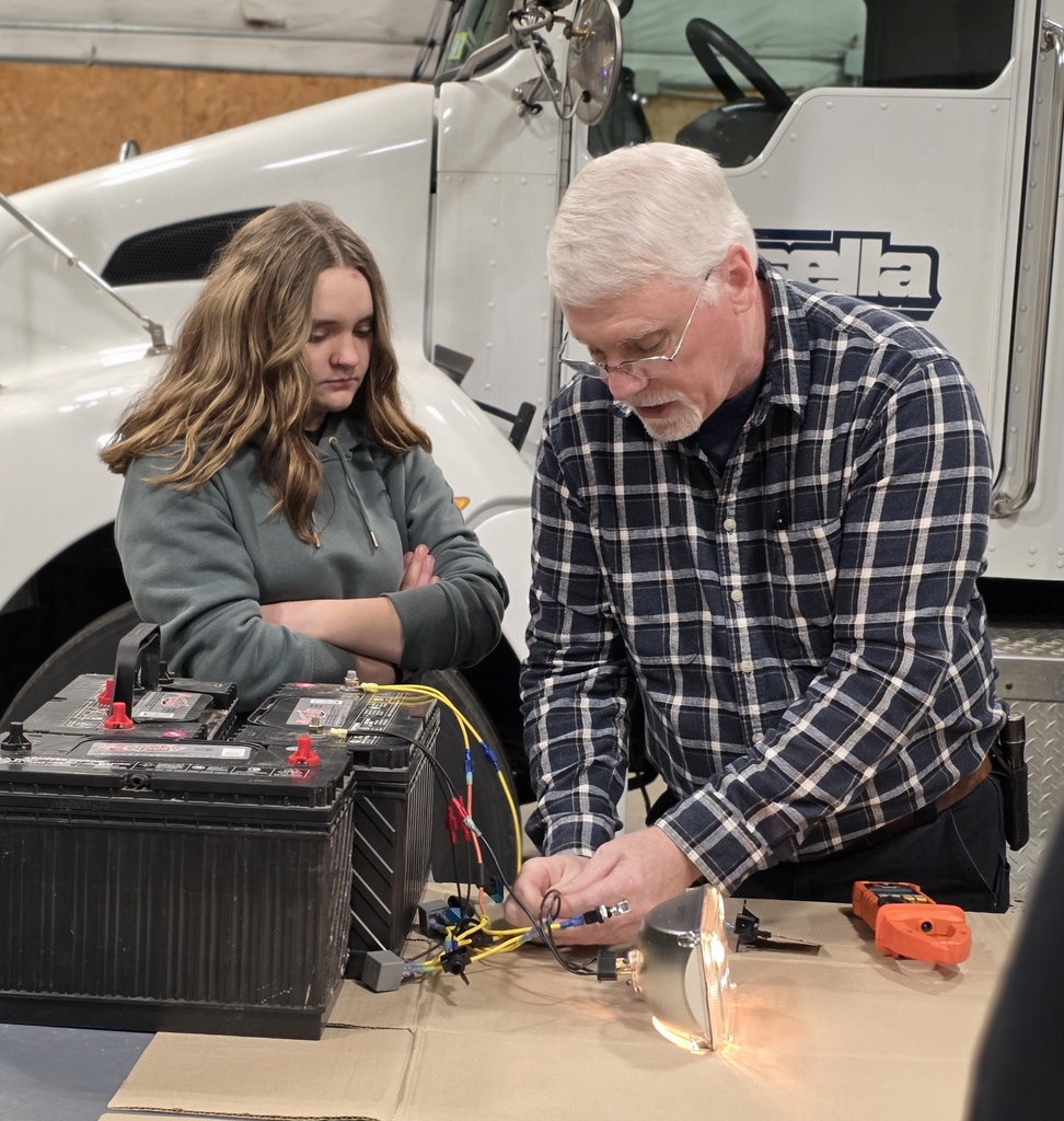 A teacher works on an electrical wiring demonstration connected to large truck batteries as a student observes closely.