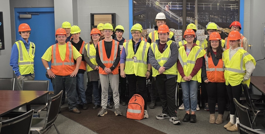 A group of high school students and staff wearing safety vests and hard hats pose together during a field trip, standing in a meeting room at Casella Waste Systems.