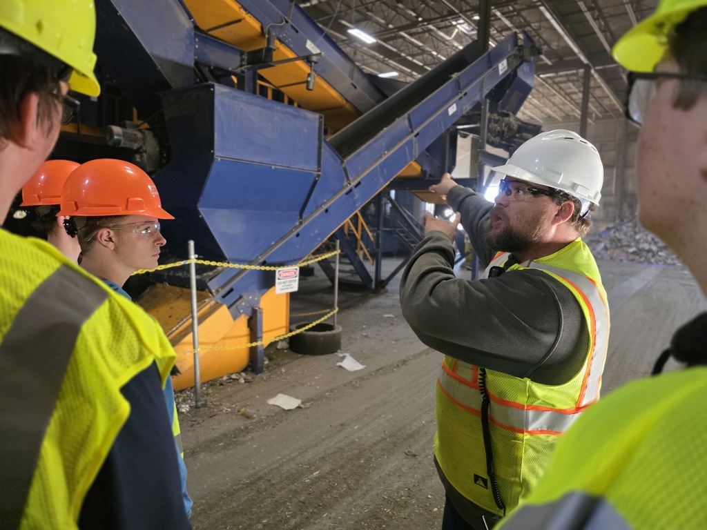 A Casella employee in a white helmet points toward a conveyor system while explaining its function to students.