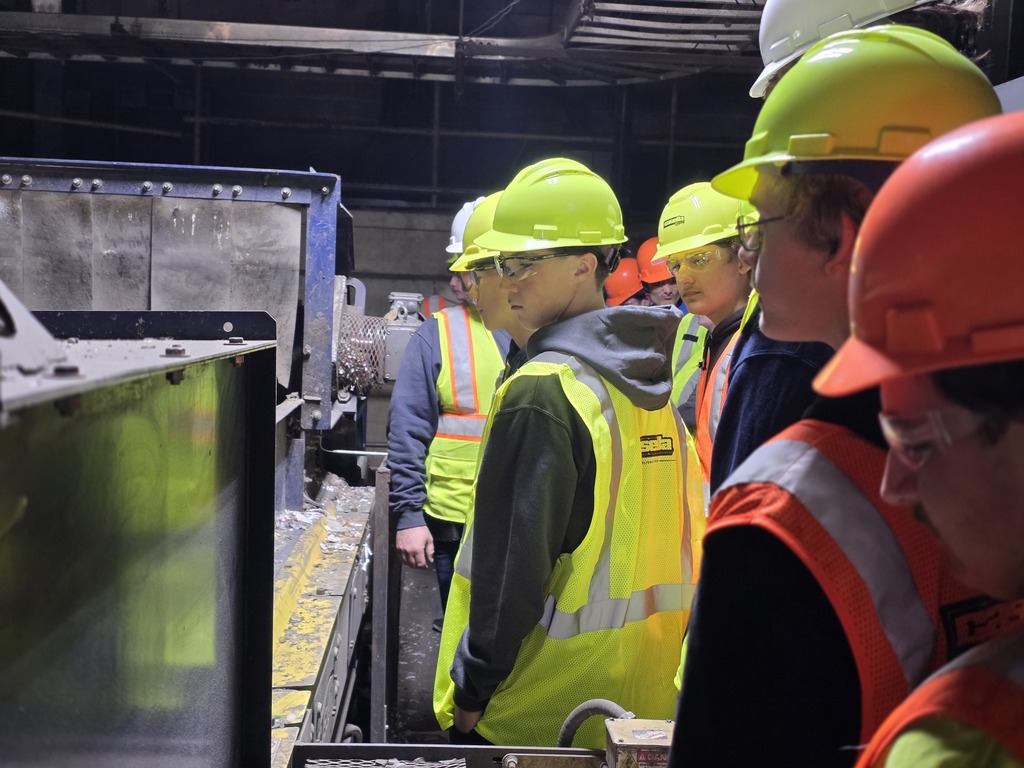 Students in bright safety gear observe a conveyor belt system in operation inside the recycling center.