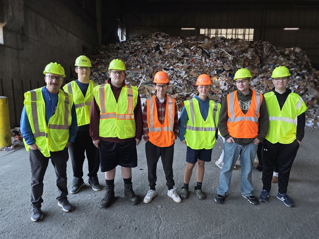 A smaller student group in reflective vests and hard hats stands in front of a large pile of recyclable materials inside the facility.