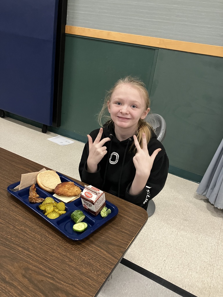 A smiling student in a black hoodie flashes peace signs while sitting at lunch with a chicken sandwich, pickles, and milk.