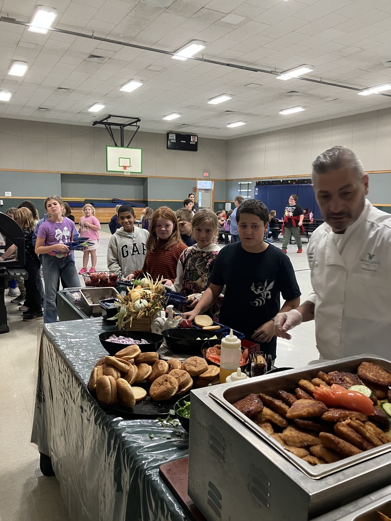 Students stand in line at a lunch buffet with buns, chicken patties, and toppings, assisted by a chef in white.