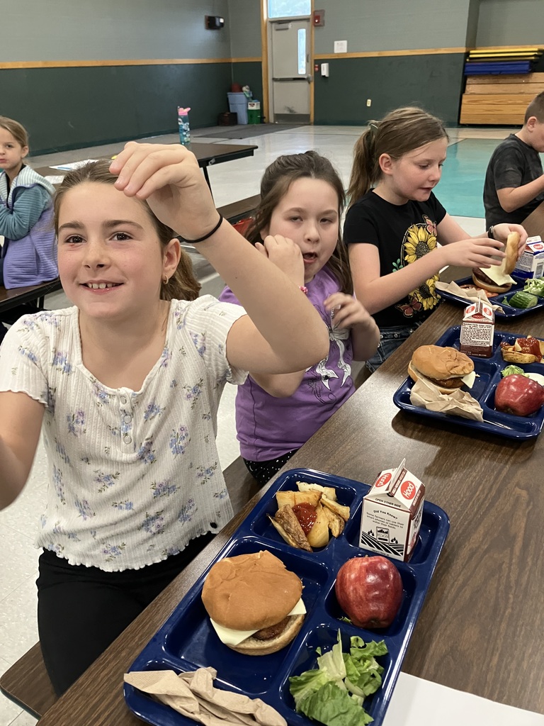 Three students sit at a lunch table smiling and eating sandwiches with fries, apples, and milk cartons.