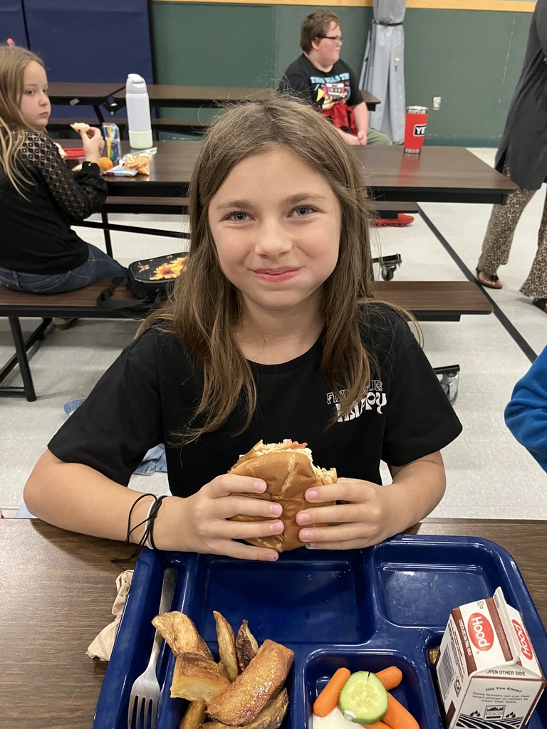A student in a black shirt smiles while holding a sandwich at the lunch table.