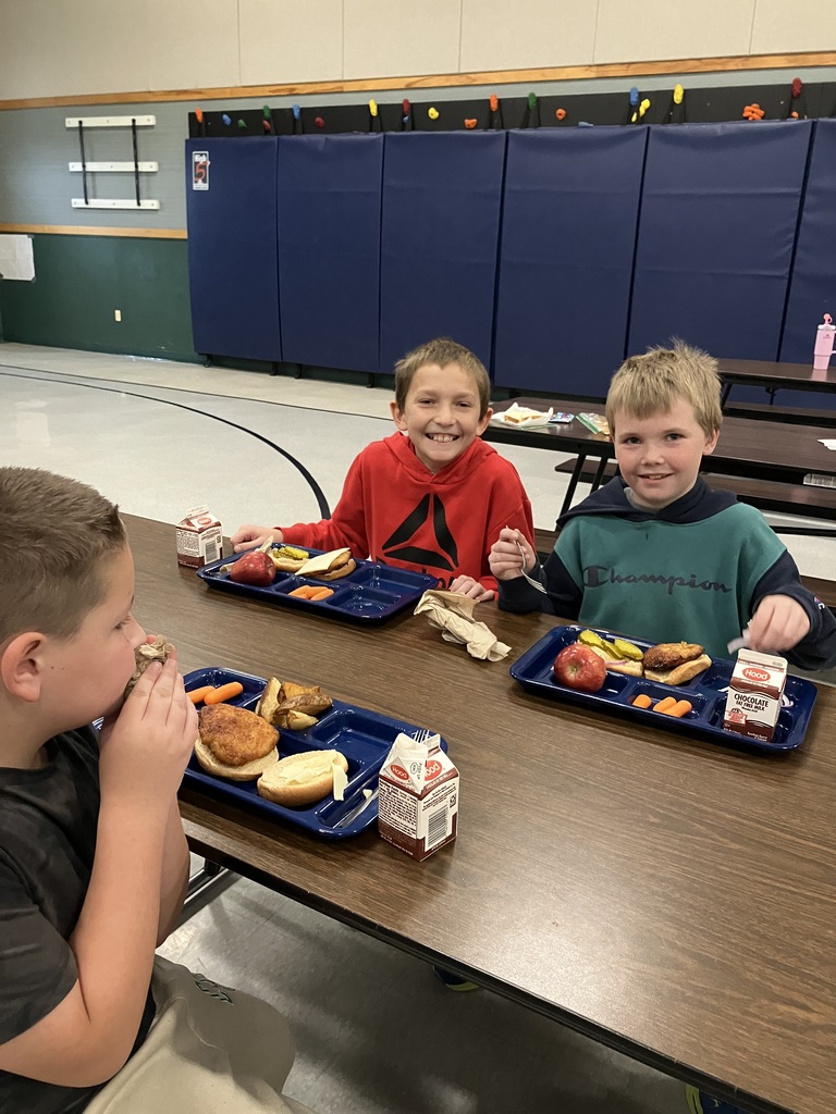 Three students smile while eating lunch together, with trays holding chicken sandwiches, carrots, apples, and milk.