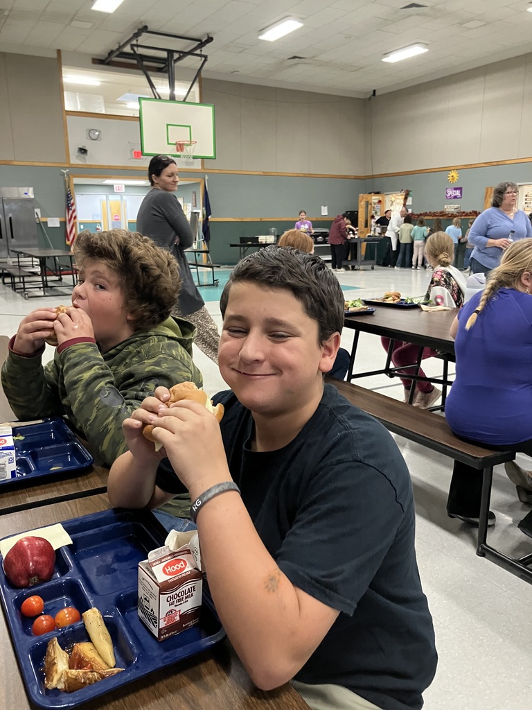 Two students sit at a cafeteria table, both eating sandwiches and smiling, with other students and staff visible in the background near the lunch line.