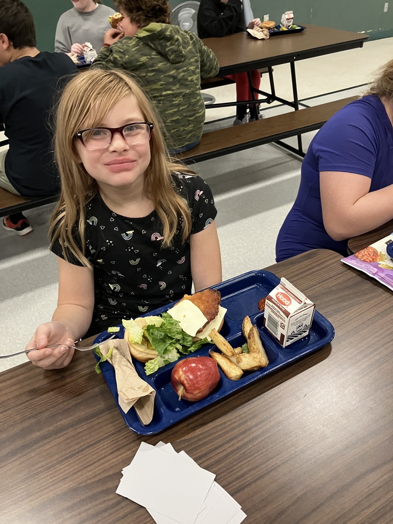 A student wearing glasses and a black shirt with rainbow hearts smiles at the camera while eating, with a tray holding a sandwich, fries, an apple, and chocolate milk.