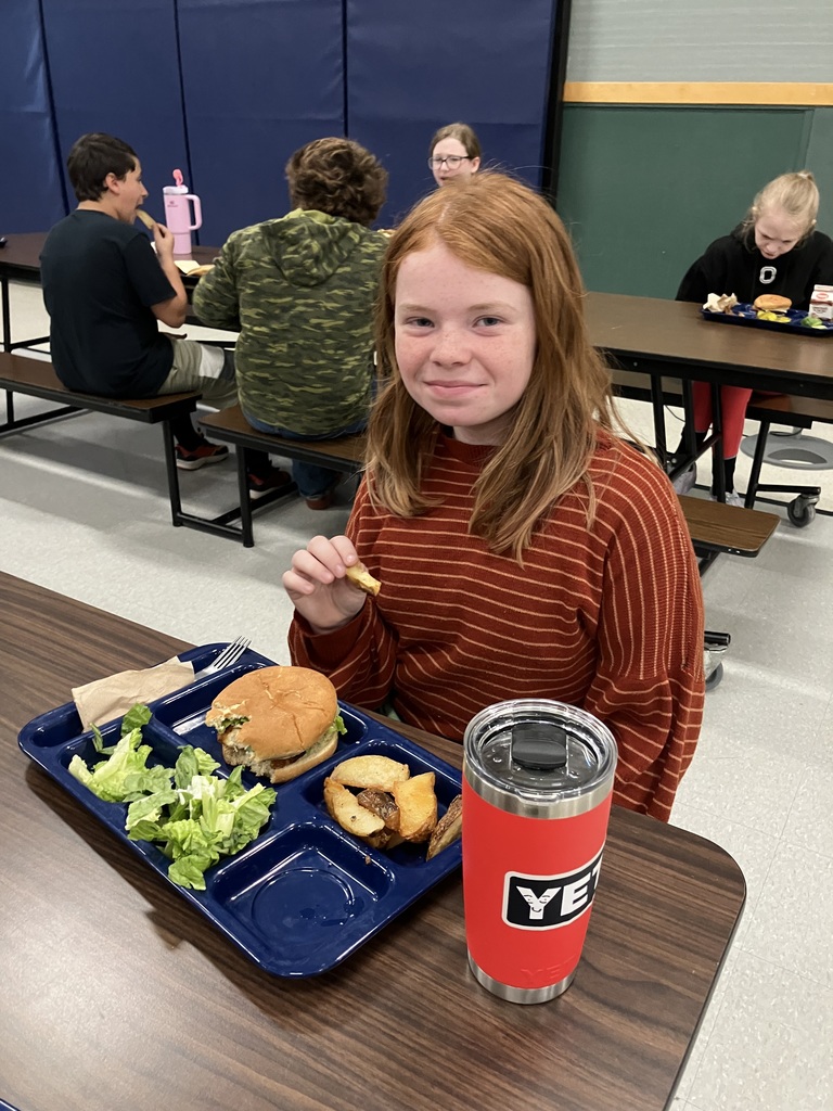 A student in a red striped sweater smiles while eating lunch, with a tray containing a sandwich, salad, and potato wedges, and a red insulated cup on the table.