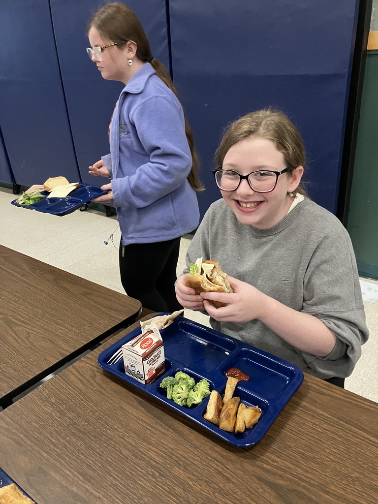 A smiling student sits at a cafeteria table holding a sandwich, with a lunch tray containing broccoli, potato wedges, and chocolate milk.