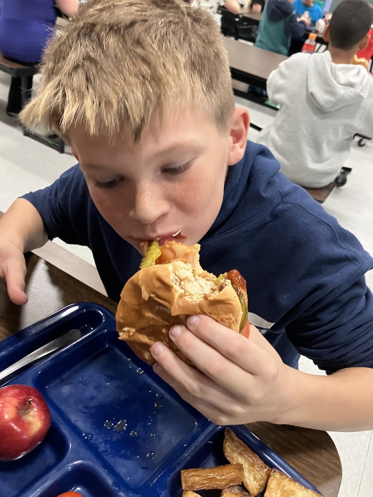 A student in a navy sweatshirt takes a big bite of a sandwich filled with lettuce, tomato, and pickles during lunch.