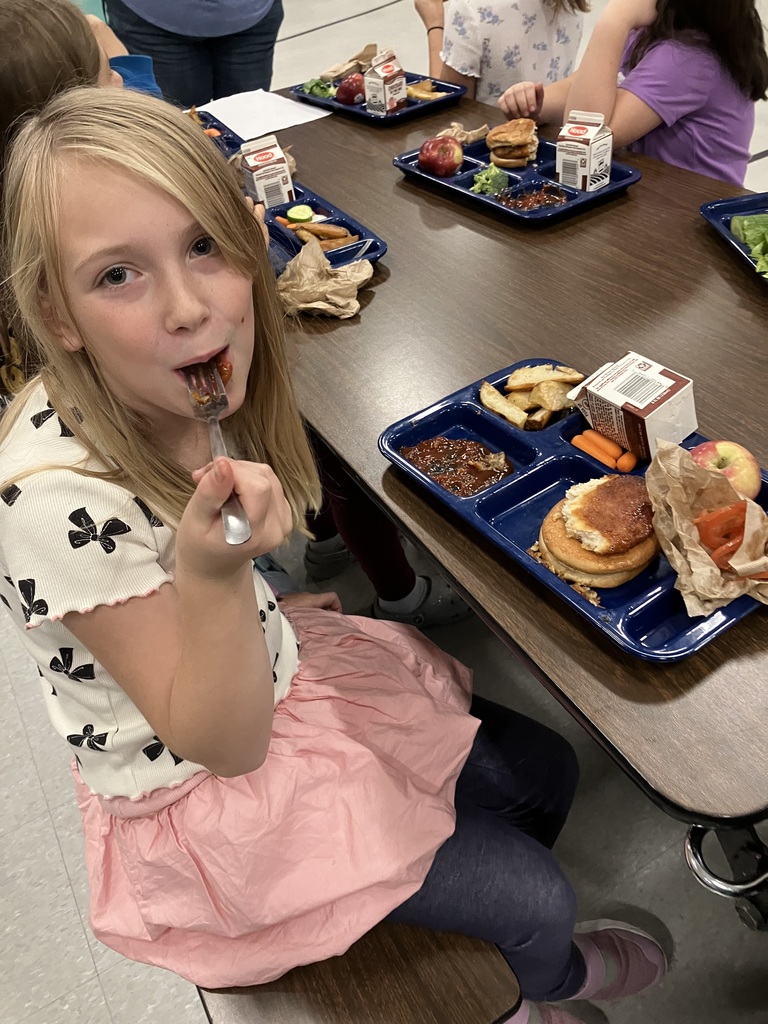 A student in a white shirt with black bows eats with a fork, with a tray containing a sandwich, carrots, potato wedges, chocolate milk, and an apple.