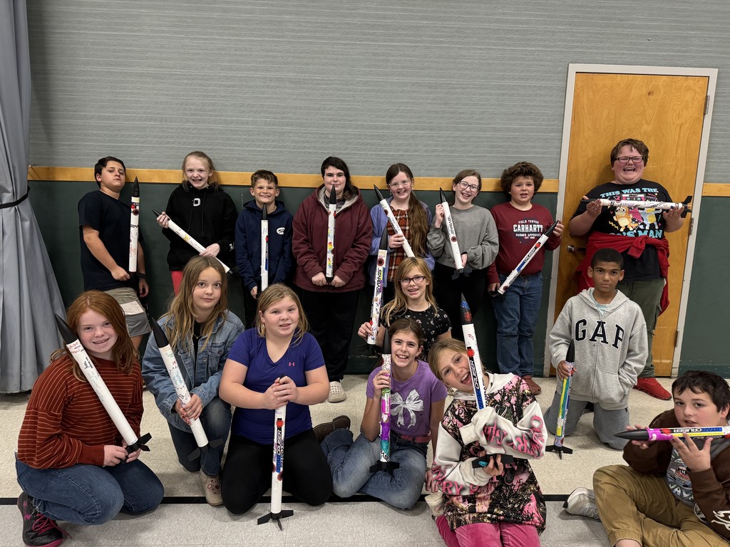 A group of elementary school students pose indoors, proudly holding decorated model rockets they built. They are smiling and kneeling or standing in rows against a gray and green wall, showing off their colorful rocket projects.