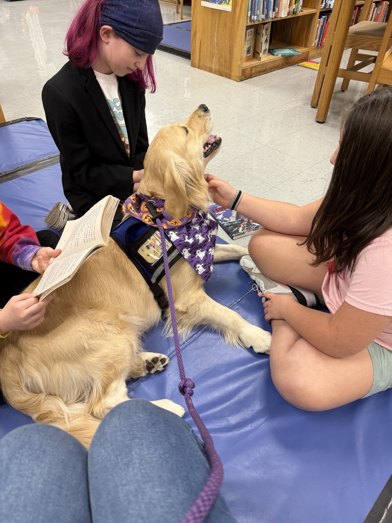 Three students sit around the golden retriever therapy dog, petting it while one student reads aloud from a book.