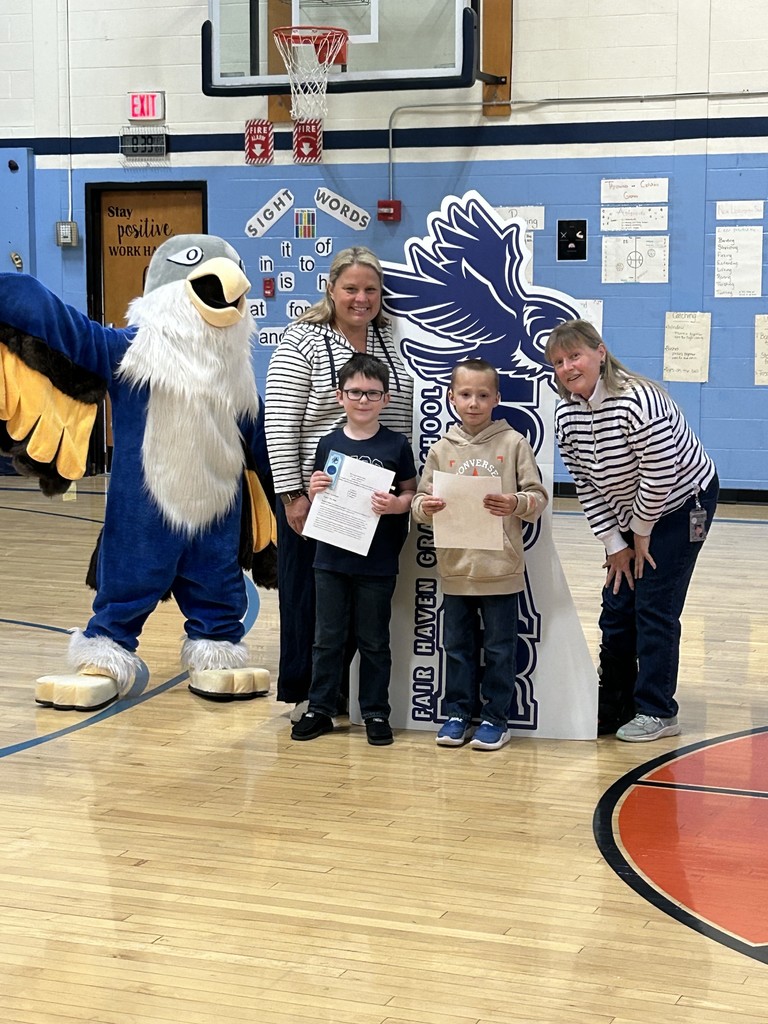 Two young boys holding papers stand with two adult women and a large blue and white bird mascot in a school gymnasium. Behind them is a cutout sign with the text "Fair Haven Grade School," a basketball hoop, and a wall with signs including "Sight Words."