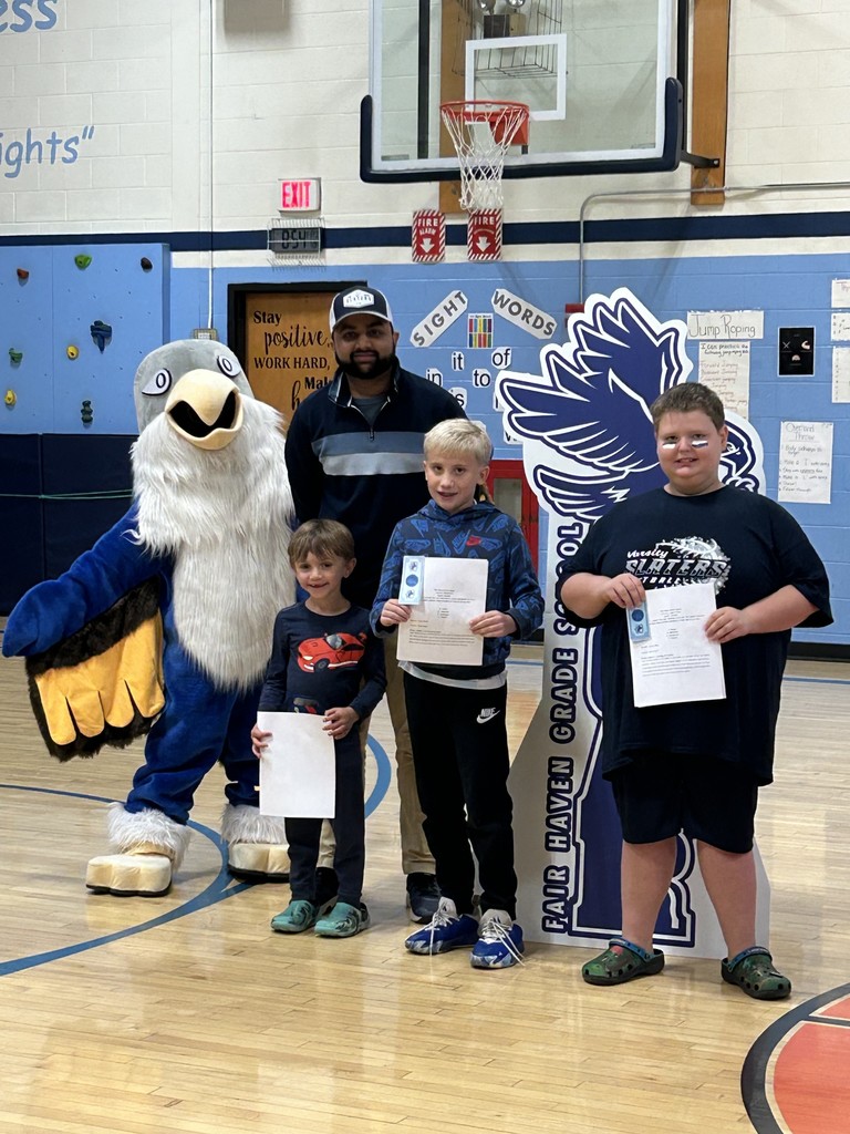 Three boys holding papers and an adult man stand with a large blue and white bird mascot in a school gymnasium. Behind them is a large, dark blue cutout sign with the text "FAIR HAVEN GRADE SCHOOL" and an image of a bird. The background includes a basketball hoop, a climbing wall, and signs for "Sight Words."