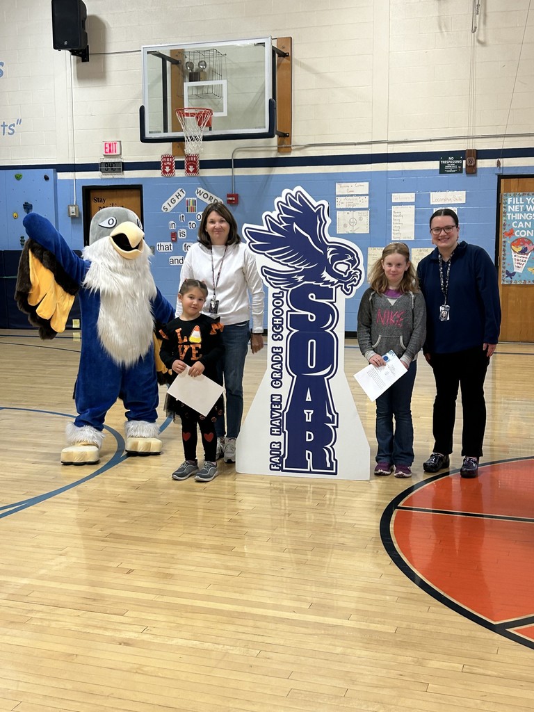 A group consisting of two young girls, two adult women, and a large blue and white bird mascot stands in a school gymnasium. The girls are holding papers. To their right is a cutout sign with the word "SOAR" next to the text "Fair Haven Grade School" and an image of a bird. The background includes a basketball hoop, a climbing wall, and signs for "Sight Words."